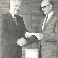 Two government officials in suits shake hands and exchange check.