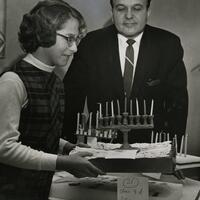 A woman places a menorah on a table near a label.  A man looks on.