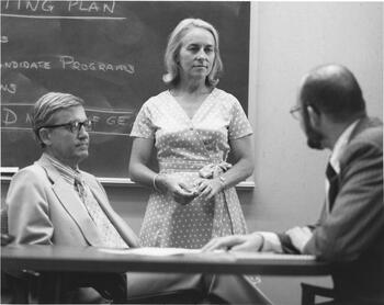 Nancy Fitzroy standing between two seated male colleagues at the GE Research Center. There is a chalkboard with writing on it behind them.  
