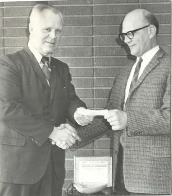 Two government officials in suits shake hands and exchange check.
